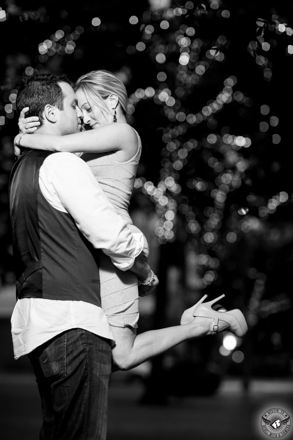 hot engagement couple embrace in front of tree with holiday lights with bokeh in background.  bride is wearing a light tight short dress  and groom is wearing an untucked white shirt with a dark vest.  she has jumped into his arms and they are hugging